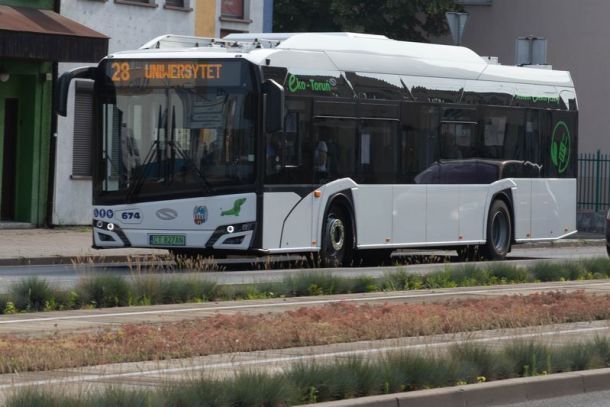 miejski autobus elektryczny koloru białego jadacy po jezdni wzdłuż torowiska tramwajowego